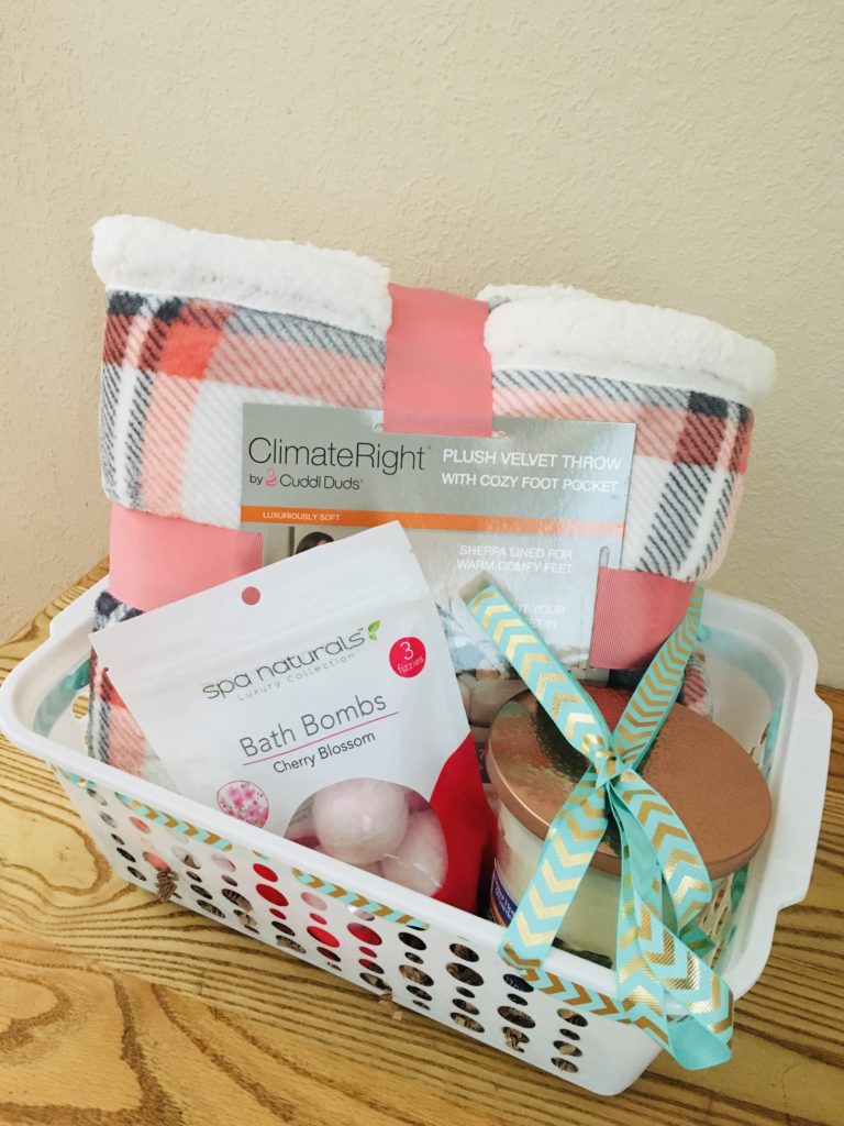 Cozy themed gift basket wrapped with ribbon that includes a large throw blanket, bath bombs, and candle 