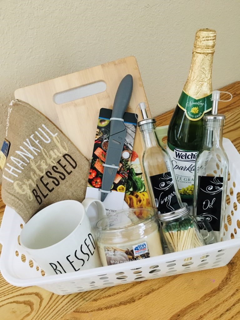 Wine gift basket with sparkling wine, cutting board, small knife, kitchen towel, vinegar and oil jars, coffee mug, candle and small jar with candle sticks